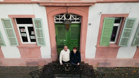 Ein älterer Mann und eine ältere Frau stehen auf der Treppe vor dem Eingang eines alten, imposanten Hauses.