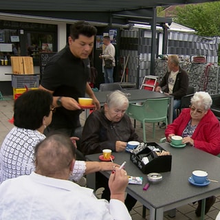 Joti trägt einen Kaffee von seinem Kiosk zu einem Tisch, an dem Seniorinnen sitzen.