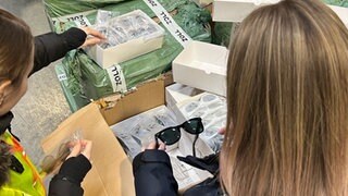 Zwei Mitarbeiterinnen des Zolls packen Sonnenbrillen aus Paketen aus. Offenbar handelt es sich dabei um Fälschungen.