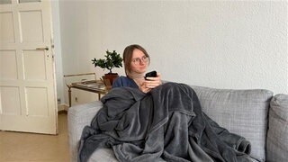 Eine junge Frau liegt eingedeckt mit dem Handy auf dem Sofa, tags: Lernen, KI, Sprache, Sprachlehrer