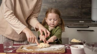 Ein Kind drückt einen Teigklumpen auf ein bemehltes Holzbrett. Dabei bekommt es Hilfe von seiner Mutter, von der nur Hände, Arme und Oberkörper zu sehen sind.