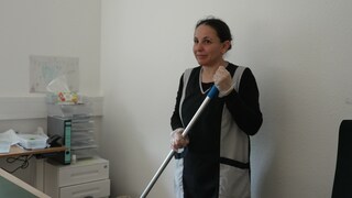 Reinigungskraft Ayse steht in einem Büro. Sie trägt einen Putzkittel und hat Handschuhe an. In den Händen hält sie einen Wischmopp.