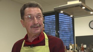 Älterer Herr mit braunen Haaren und Brille steht mit einer grünen Schürze und einem roten Strickpulli in einer Küche.