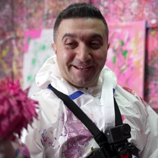 Ein Mann steht in einem Raum, in dem alle Wände voll sind von Farbspritzern. Der Mann trägt einen Schutzanzug und hält einen mit Farbe getränkten Ball in der Hand. 