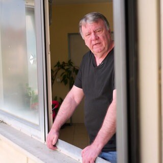 Dieter Scheibel steht an einem offenen Fenster und hält sich mit beiden Händen an der Fensterfassung fest. Er schaut Richtung Kamera. Die Fassade des Hauses ist leicht gelblich.