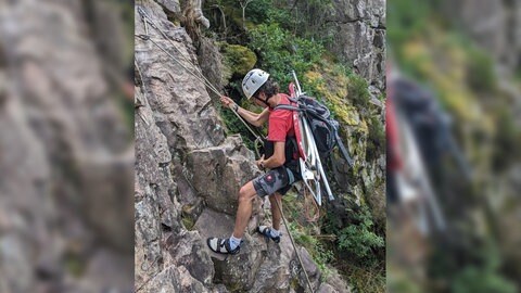 Extrembügler klettert eine Felswand mit dem Bügelbrett auf dem Rücken hoch.