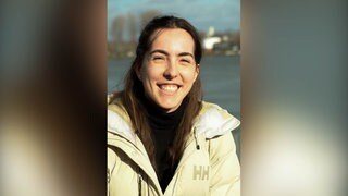 Junge Frau mit dunklen Haaren und beiger Jacke lächelt in Kamera.
