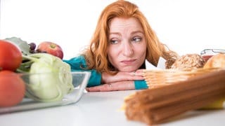 Eine Frau stützt Arme und Kopf auf einen Tisch und schaut missmutig auf eine Schale mit Ost und Gemüse: Wer raucht, sich nicht viel bewegt und schlecht ernährt, erhöht sein Risiko für viele schwere Erkrankungen. 
