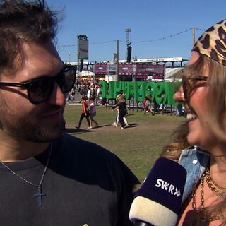 Jannik und Patricia sind auf dem Glückgefühle Festival auf dem Hockenheimring. Sie tragen Sonnenbrillen und schauen sich an.