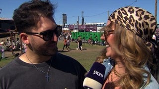 Jannik und Patricia sind auf dem Glückgefühle Festival auf dem Hockenheimring. Sie tragen Sonnenbrillen und schauen sich an.