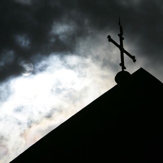 Im Gegenlicht und vor wolkenverhangenem Himmel ist die Kirchturmspitze eines Doms mit Kreuz zu sehen.