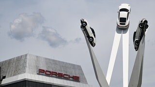 Der Schriftzug von Porsche ist an einem Gebäude am Stammsitz angebracht. Rechts daneben sind Porsche-Fahrzeuge an einem Kunstwerk auf dem Porscheplatz angebracht.