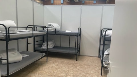 Drei Stockbetten aus dunkelgrauen Stahlrohren stehen eng nebeneinander in einer Kabine aus dünnen Messewende - so werden Geflüchtete in einer Sporthalle in Friedrichshafen untergebracht