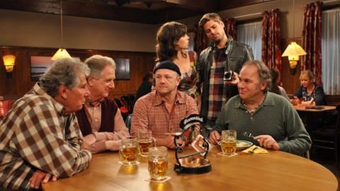 Leo, Schorsch, Toni und Ludwig sitzen am Stammtisch, dahinter stehen Eva und Andreas