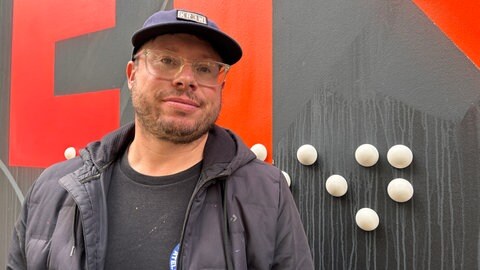 Der französische Künstler THE BLIND steht vor einer schwarz-roten Wand mit einem Relief in Braille-Schrift