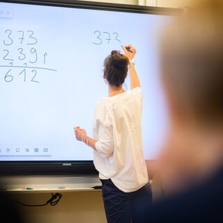 Eine Referendarin schreibt eine Mathematikaufgabe auf eine digitale Schultafel in einem Klassenraum. 
