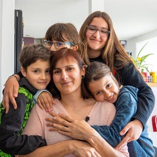 Frau in der Mitte des Bildes wird von vier Kindern von hinten umarmt.