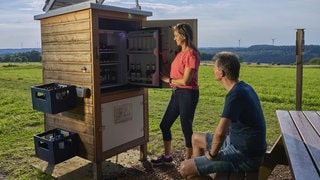 Eine Frau steht vor einer geöffneten Getränkestation auf der Wandertour "Lecker Pfädchen", daneben sitzt ein Mann auf einer Bank.