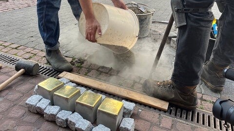Menschen stehen auf einem Bürgersteig vor einem Haus und tragen Texte vor, währenddessen werden goldfarbene Steine im Boden verlegt. Vier der insgesamt 20 Stolpersteine sind für Familie Allmayer. 