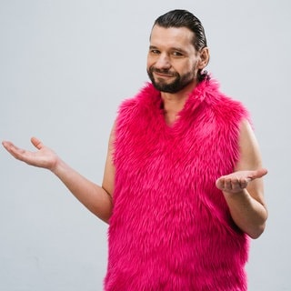 Martin Puntigam, Kabarettist und Schauspieler, dreht in pinker Puschelweste die Handflächen nach oben. Er kombiniert Topwissenschaft und Spitzenhumor.