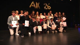 Die Abschlussklasse der Oskar-Paret-Schule steht auf einer Bühne, alle gekleidet in Anzügen oder Kleidern. Sie halten Zeugnismappen in der Hand. Im Hintegrund steht "AK26" in Ballons.