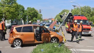 Ein orangefabenes Auto steht an einer Kreuzung, umgeben von Rettungskräften. Vor dem Auto ist ein umgekippter Ampelmast zu sehen. Er hat eine Fußgängerin bei einem Autounfall schwer verletzt.