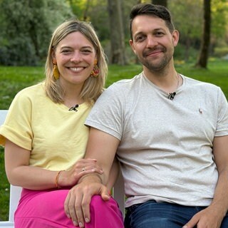 Janina und Bastian sitzen auf einer Bank und lächeln in die Kamera. Im Hintergrund ist ein rot-gelber Ballon zu sehen.