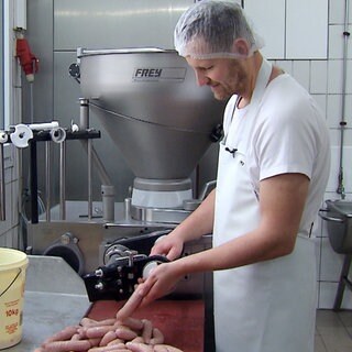 Philipp Burkhardt trägt weiße Arbeitskleidung und eine durchsichtige Haube. In der Hand lässt er mehrere rohe Weißwürste durch seine Hände gleiten.