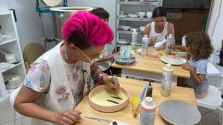 Eine Frau mit pinkfarbenem Haar beugt sich über einen Teller und bemalt ihn. Auch Melanie Traunig aus Jedelhausen bei Neu-Ulm folgt dem Trend, Keramik selbst zu bemalen und schafft sich so ihr eigenes, buntes Geschirr. 