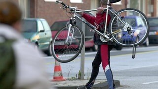 Mann trägt Fahrrad über die Straße. Immer mehr Menschen sind mit dem Rad unterwegs. Was auf dem Fahrradweg oder an Ampeln erlaubt ist. Wann es für Radfahrer Punkte in Flensburg gibt. Was beim Zubehör wichtig ist.