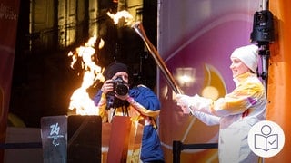 Eine Sportlerin entzündet die olympische Fackel an einer Feuer·stelle. Im Hintergrund macht ein Fotograf ein Foto. Rechts das SWR Logo für Leichte Sprache. 