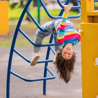Kleines Mädchen hängt kopfüber an einem Klettergerüst: Bei Spielplätzen prallen Interessen aufeinander. Kinder wollen spielen, Eltern Sicherheit, Kommunen DIN-Normen einhalten und die sinkenden Budgets abfedern. 