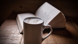 Symbolbild: Eine Tasse Kaffee mit einem aufgeschlagenen Buch.