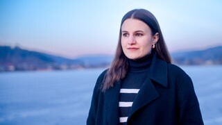 Die ehrenamtliche Sterbebegleiterin Katharina steht vor einem See. Sie trägt ihre Haare offen und hat einen schwarzen Mantel an, unter dem sie einen schwarz-weißen Rollkragenpullover trägt. 