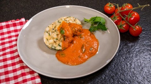 Auf einem hellgrauen Teller sind eine Scheibe Serviettenknödel mit einer Tomatensauce angerichtet und mit Petersilie dekoriert. Daneben liegt ein Bund Cherrytomaten und eine rot-weiß-karierte Stoffserviette.
