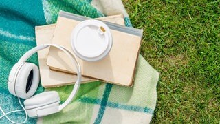 Bücher, Kopfhörer und Kaffee liegen auf einer Picknickdecke in einer Wiese