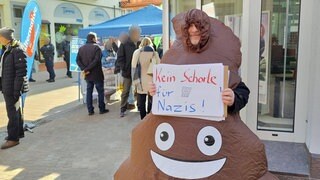 Mann protestiert in Neustadt in einem Kostüm, das wie ein Kackhaufen aussieht