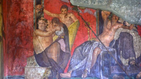 Fresko des Dionysos-Kults aus der Villa der Mysterien in Pompeji mit rotem Hintergrund und mehreren Figuren, die an rituellen Handlungen teilnehmen.