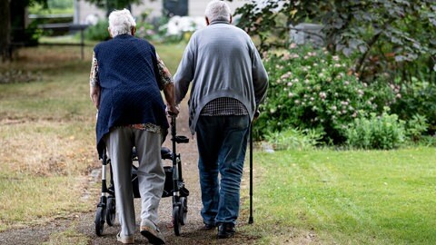 Eine Seniorin mit Rollator und ein Mann mit Stock gehen spazieren. Symbolbild für die Debatte über gestiegene Pflegekosten in Rheinland-Pfalz