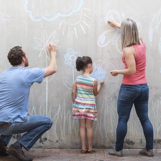 Zwei Erwachsene und ein Kind bemalen eine Wand mit Kreide