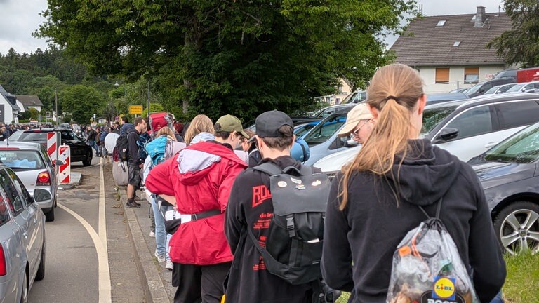 Mehrere Festivalbesucher stehen mit Rucksäcken und Campingausrüstung in einer Schlange am Straßenrand.