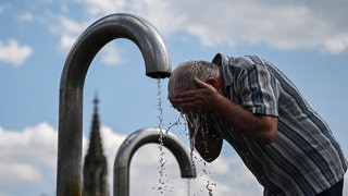 Ein Passant kühlt sich an einem Mineralwasserbrunnen ab, im Hintergrund ein Kirchenturm.