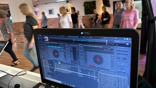 Ein Monitor zeigt abgespielte Musik, im Hintergrund tanzen Menschen