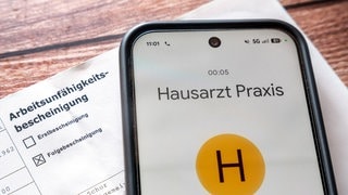Ein Smartphone mit Anruf beim Hausarzt liegt neben einer Arbeitsunfähigkeitsbescheinigung: Die Bundesregierung plant eine Krankschreibung ab dem ersten Tag (neue Reform) und das Ende der telefonischen Krankschreibung.