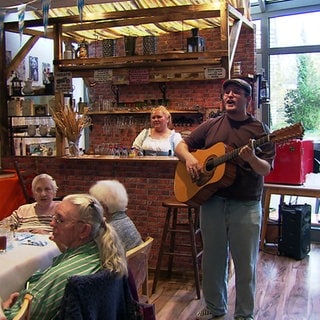 Kneipe in einem Pflegeheim in Ilvesheim: Im Vordergrund sitzen Bewohner an einem Tisch, ein Mann mit Gitarre steht im Raum und singt, im Hintergrund ist ein Tresen zu sehen. 