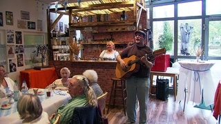 Kneipe in einem Pflegeheim in Ilvesheim: Im Vordergrund sitzen Bewohner an einem Tisch, ein Mann mit Gitarre steht im Raum und singt, im Hintergrund ist ein Tresen zu sehen. 