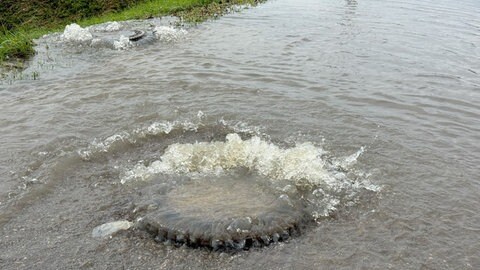 Aus Kanaldeckeln auf einer Straße sprudelt Wasser. Bei einem Gewitter mit Starkregen am Sonntagnachmittag ist die Kanalisation in Dornstadt (Alb-Donau-Kreis) überfordert. Die Wassermassen drücken Kanaldeckel hoch. Die Autobahn 8 ist kurzzeitig überflutet und muss für eine halbe Stunde gesperrt werden.