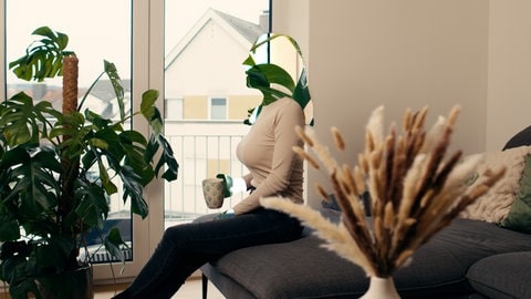 Katrin sitzt mit einer Tasse in der Hand auf einem Sofa. Sie schaut geradeaus. Ihr Gesicht und ihre Hände sind unkenntlich gemacht.