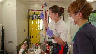 Eine Notfallanitäterin und eine Notärztin stehen in einem Krankenwagen und kümmern sich um eine Patientin.