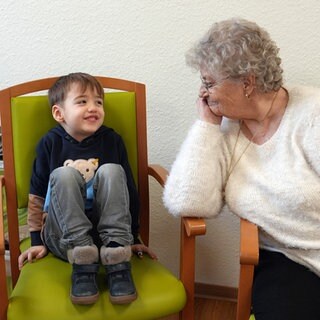 Eine alte Dame in weißem Pullover schaut einem kleinen Jungen zu, wie er kichernd auf seinem Stuhl sitzt.
